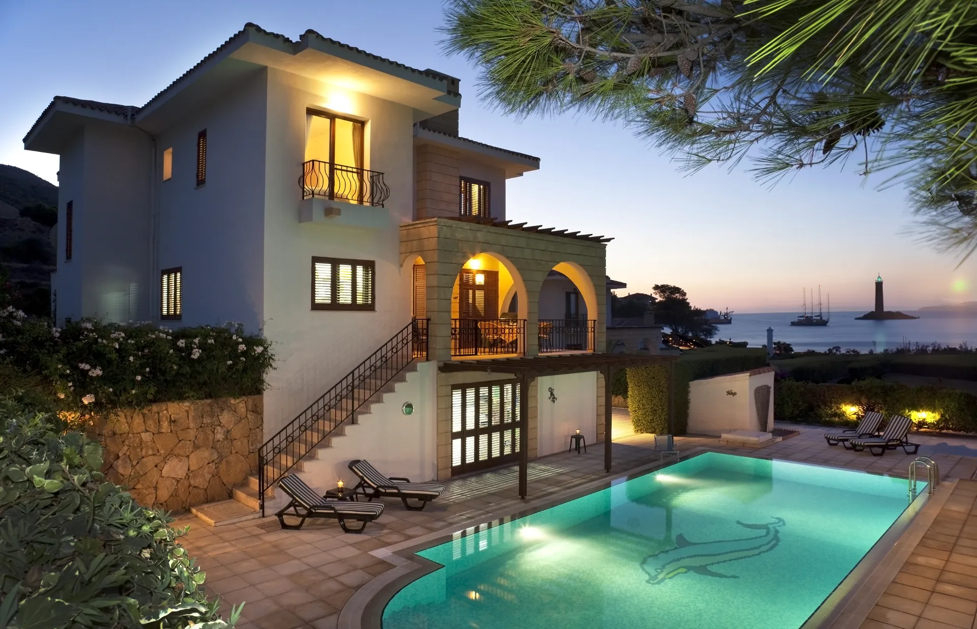 Villa terraces stepping down to a lit pool at sunset by the sea